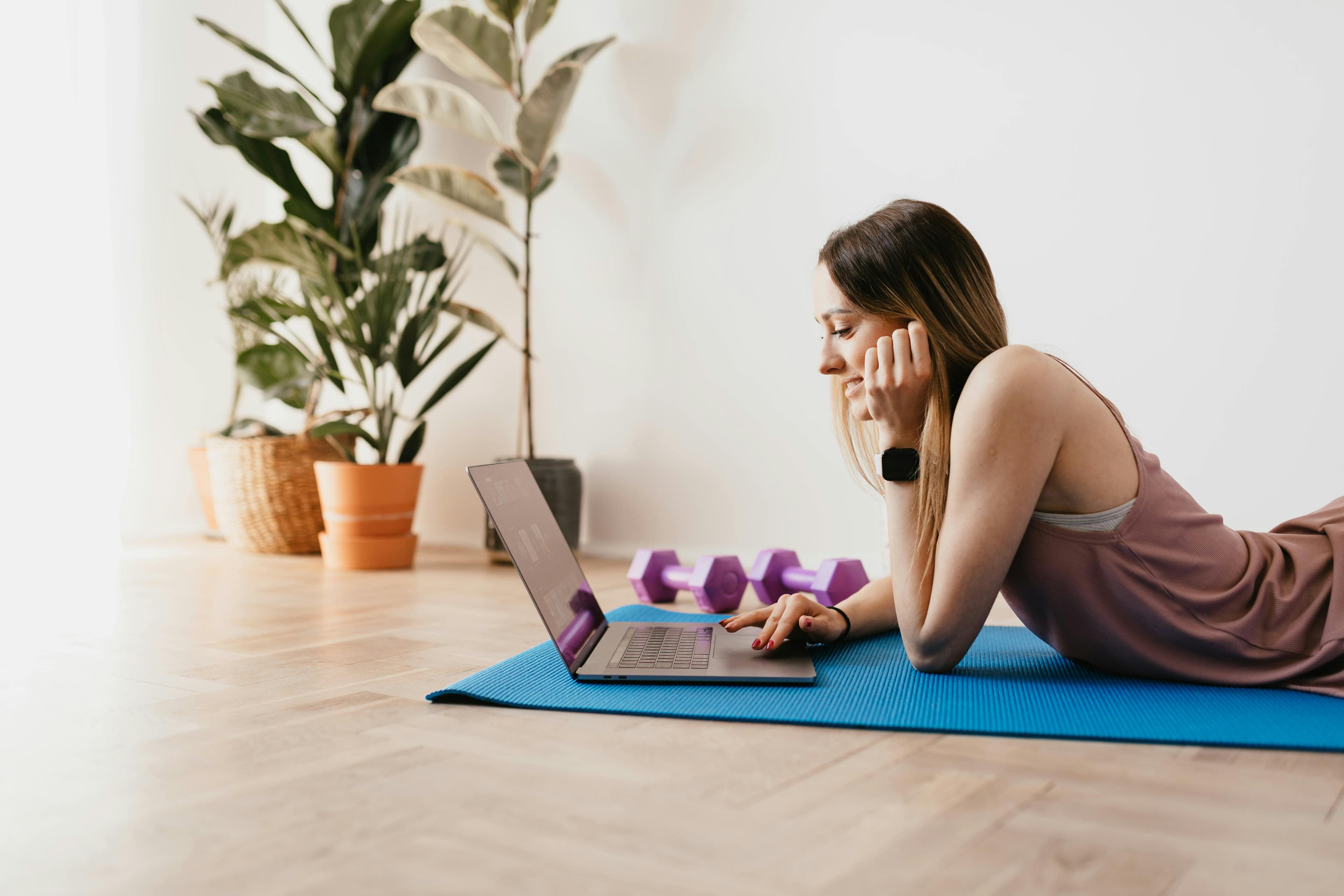 Woman on a yoga mat with dumbbells, smiling at her laptop during a workout session